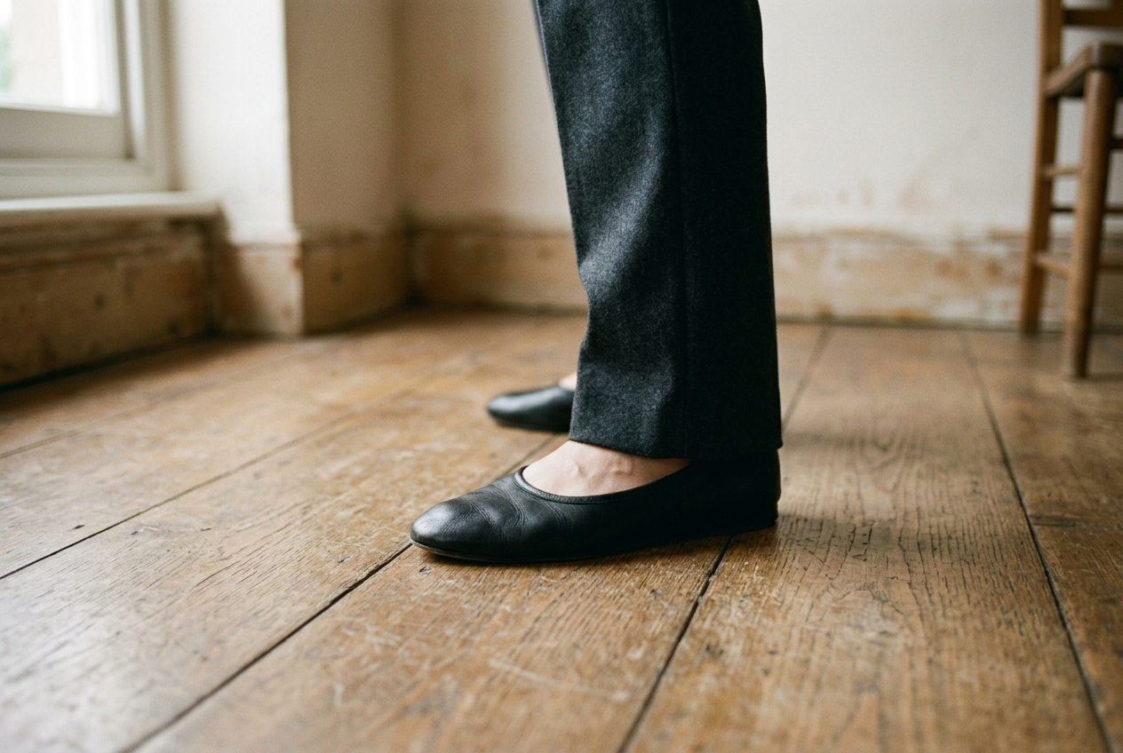 Black leather almond-toe ballet flat beneath the hem of straight charcoal trousers.