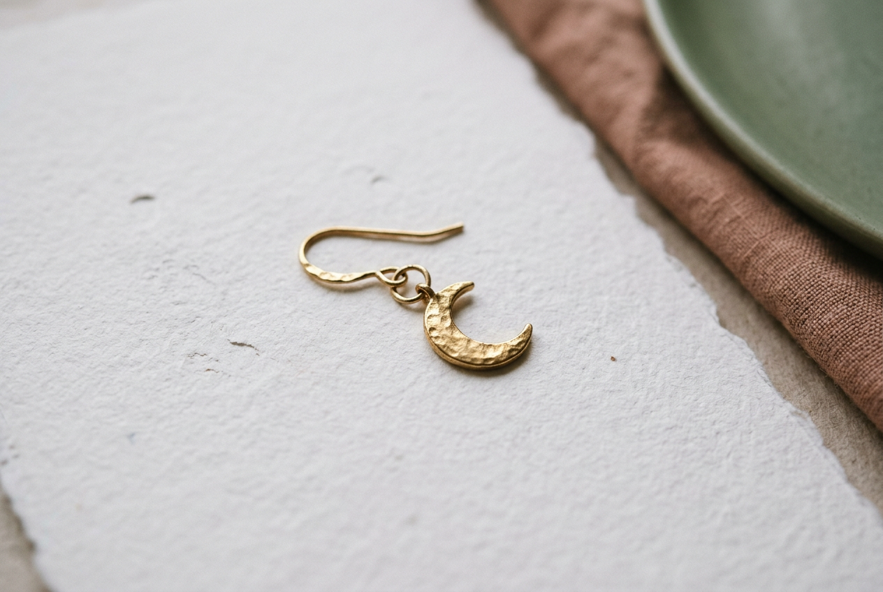 Close view of a gold-tone crescent earring showing its hook, small jump ring, and flat charm on white paper.