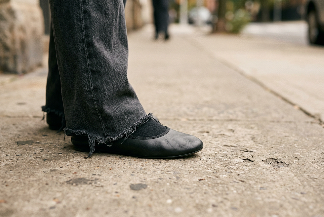 Black rounded ballet flat beneath the hem of charcoal wide-leg trousers on a plain sidewalk.